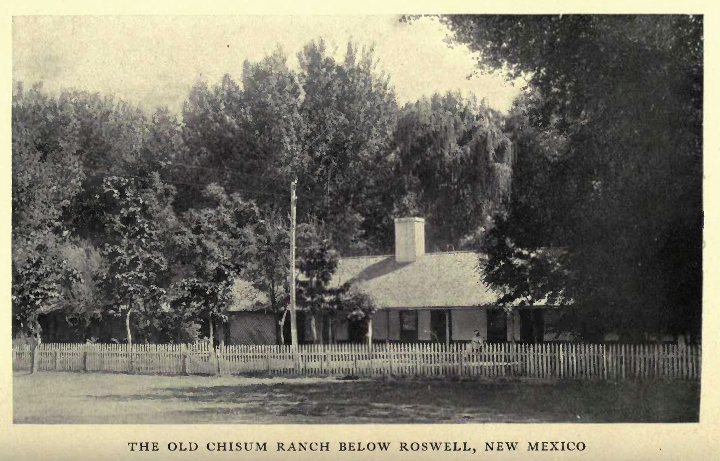 A long low ranch house behind a picket fence, framed by cottonwoods